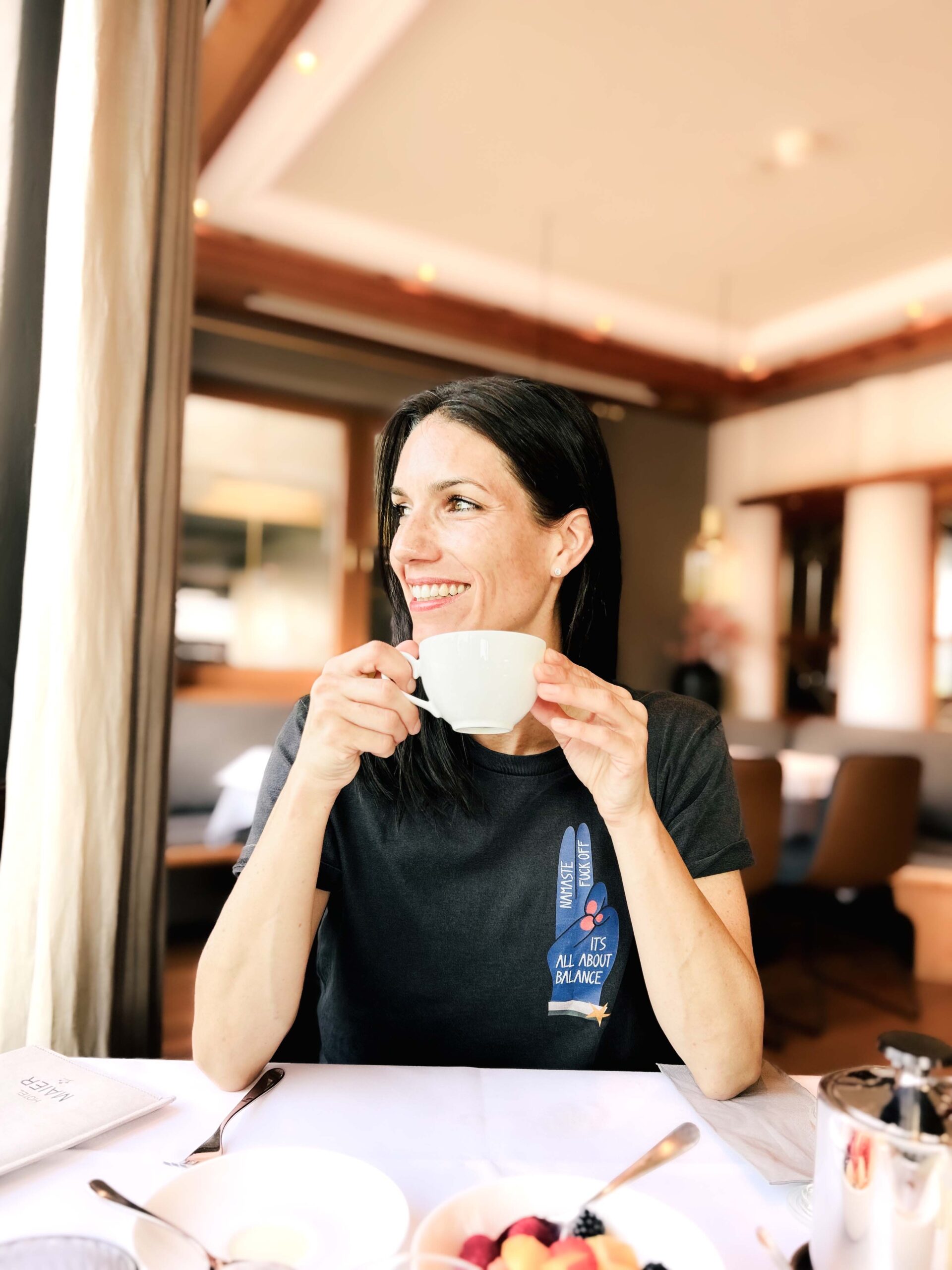 Eine Frau mit dunklen Haaren sitzt in einem hellen Restaurant am Tisch. Vor ihr stehen ein Teller mit frischem Obst, Besteck und eine Kanne. Sie trägt ein dunkles T-Shirt mit Aufdruck und hält eine weiße Tasse in beiden Händen. Mit einem freundlichen Lächeln schaut sie aus dem Fenster ins Licht.